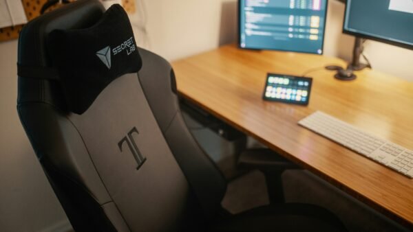 Silla gaming negra junto a un escritorio de madera con doble monitor y setup de trabajo moderno.